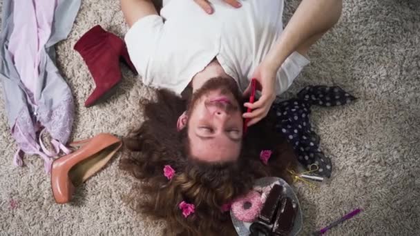 Краєвид щасливої кавказької інтерсексуальної людини лежить на м'якому килимі і розмовляє по телефону. Портрет бінарного гендерного чоловіка з гримом на обличчі і квітами в кучерявому волоссі, оточеного жіночими речами.. — стокове відео