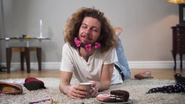 Portrait de jeune homme caucasien souriant avec des fleurs à la barbe couché sur un tapis doux et buvant du thé ou du café. Intersexualité positive exprimant l'individualité. Genre binaire, perception de soi . — Video