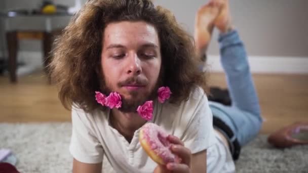 Close-up van positieve Kaukasische intersekse persoon liggend op de vloer en smakelijke donut etend. gelukkig man vrouw met bruine ogen en bloemen in baard kijken naar camera en glimlachen. — Stockvideo