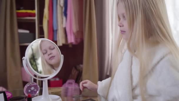 Vista lateral da menina branca bonita refletindo no espelho. Criança loira bonito em roupão de banho branco em casa. Moda, infância, estilo de vida . — Vídeo de Stock