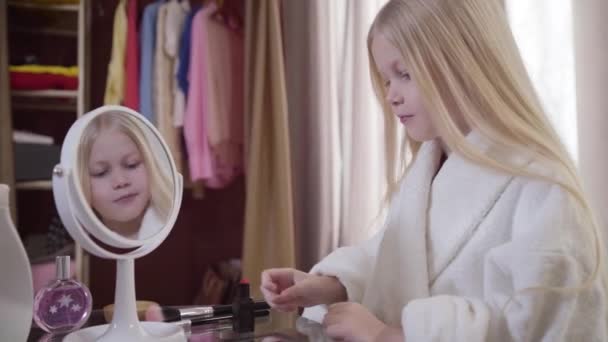 Camera approaching to face of excited blond Caucasian child taking red lipstick from the table. Charming girl choosing cosmetics indoors. Fashion, style, beauty. — 비디오