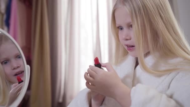 Close-up de menina branca loira abrindo batom vermelho e balançando a cabeça. Criança bonita elegante em roupão branco escolhendo cosméticos em casa. Beleza, cuidado, estilo de vida . — Vídeo de Stock