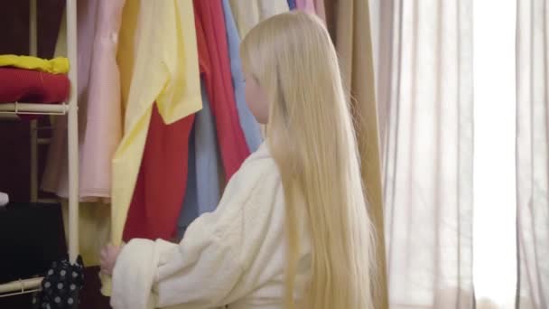 Schattig klein meisje met lang blond haar en grijze ogen die naar garderobe komen, naar kleren kijken en denken. Mooie kindkeuze outfit in de ochtend. Stijl, mode, kindertijd. — Stockvideo