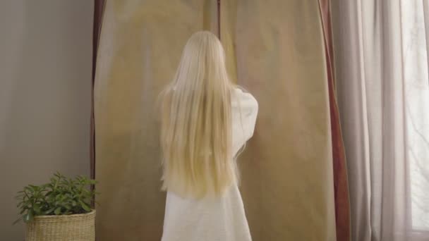 Achteraanzicht van het kleine blanke meisje met lang blond haar dat naar de kast komt en het opent. Vrolijke jonge dame selecteren jurk in de ochtend binnen. Mode, stijl, levensstijl. — Stockvideo