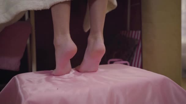 Primer plano de las niñas caucásicas pies de pie sobre los dedos de los pies. Niña en albornoz blanco de pie en la cama de color rosa y tratando de llegar a la parte superior del armario. Moda, estilo, infancia . — Vídeos de Stock