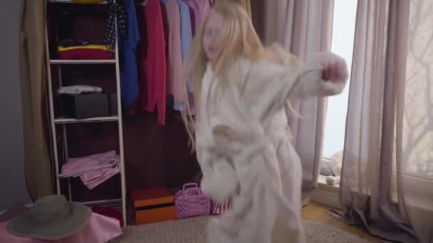 Danza energica di allegra bambina caucasica vestita con accappatoio bianco. Carino bambino biondo che salta nella sua stanza di fronte all'armadio. Gioia, divertimento, svago . — Video Stock