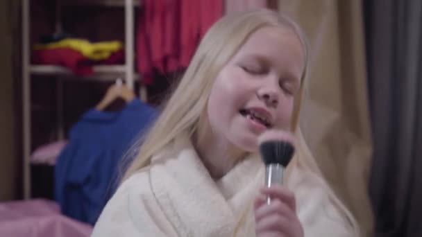 Primo piano della bella ragazza caucasica bionda che canta usando il pennello per il trucco come microfono. Bambino positivo in accappatoio bianco a riposo a casa. Hobby, tempo libero, felicità . — Video Stock