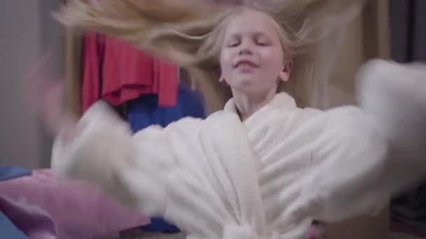 Portrait of joyful Caucasian girl shaking long blond hair and looking at camera. Happy kid in white bathrobe posing indoors. Happiness, leisure, lifestyle. — Stock Video