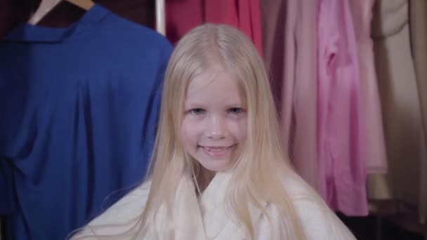 Cara de cerca de la hermosa chica caucásica con los ojos grises y el pelo largo y rubio mirando a la cámara y sonriendo. Retrato de un niño encantador en albornoz blanco posando en casa . — Vídeo de stock