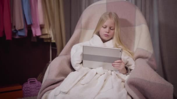 リラックスした白人ブロンドの女の子の肖像画は、自宅で肘掛け椅子に座ってタブレットを使用しています。デバイス画面を見て驚くような顔をするかなり小さな女性。インターネット、ソーシャルメディア、ライフスタイル. — ストック動画