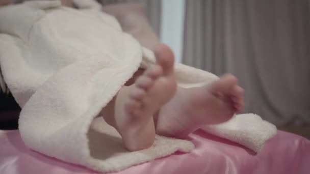 Zbliżenie stóp małej białej dziewczynki w białym szlafroku leżącej na różowym łóżku. Dziecko odpoczywa po prysznicu w swoim pokoju. Styl życia, wypoczynek, relaks. — Wideo stockowe