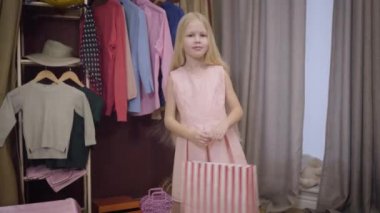 Pembe elbiseli neşeli, beyaz bir kızın portresi. Alışveriş çantasıyla evde duruyor. Mutlu çocuk pakete bakıyor ve gülümsüyor. Yaşam tarzı, moda, eğlence..
