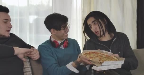 Three Caucasian male adult students sitting on couch and eating pizza. Boys talking and laughing indoors. Men having fun. Cinema 4k ProRes HQ. — Stock Video