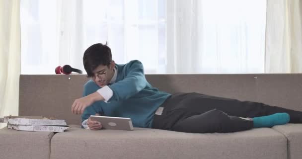 Gözlüklü mutlu beyaz adamın portresi kanepeye uzanmış tablet kullanıyor ve bir dilim pizza alıyor. İçeride dinlenen neşeli yetişkin öğrenci. Sinema 4k Prores Merkezi. — Stok video