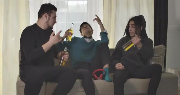 Három fiatal kaukázusi férfi ül a kanapén sörrel és beszélgetnek. Felnőtt egyetemisták lógnak otthon. Mozi 4k Prores Hq. — Stock videók