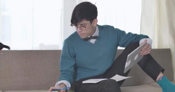 Portrait d'un étudiant universitaire caucasien intelligent en lunettes assis avec une tablette sur le canapé et regardant à travers ses notes. Beau jeune homme brune qui fait ses devoirs à l'intérieur. Siège social Cinema 4k ProRes . — Video