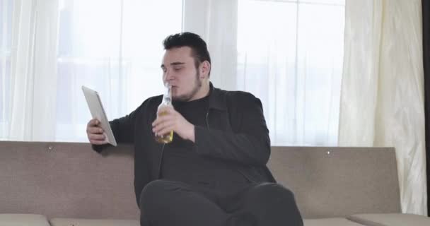 Retrato de un joven caucásico relajado bebiendo cerveza y usando tableta. Un tipo regordete descansando en casa los fines de semana. Sede del cine 4k ProRes . — Vídeos de Stock