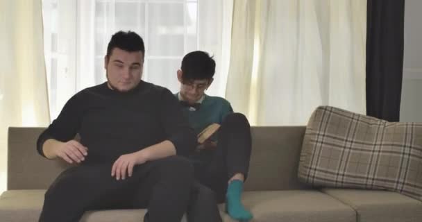 Annoying roommates coming to focused Caucasian boy reading and sitting down on couch on both sides. University students bullying nerd guy in eyeglasses. Cinema 4k ProRes HQ. — Stock Video