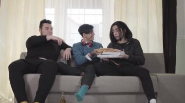 Pizza yiyen ve içeride bira içen üç beyaz genç adam. Erkek üniversite öğrencileri kanepede oturup konuşuyorlar. Neşeli ev arkadaşları evde parti yapıyor. Yaşam tarzı, rahatlama, rahatlama..