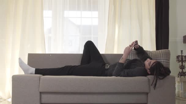 Юный кавказский мальчик с дредами, лежащий на диване, как его толстый друг входит в комнату и толкает его с дивана, чтобы сесть. Портрет двух веселых парней в помещении . — стоковое видео