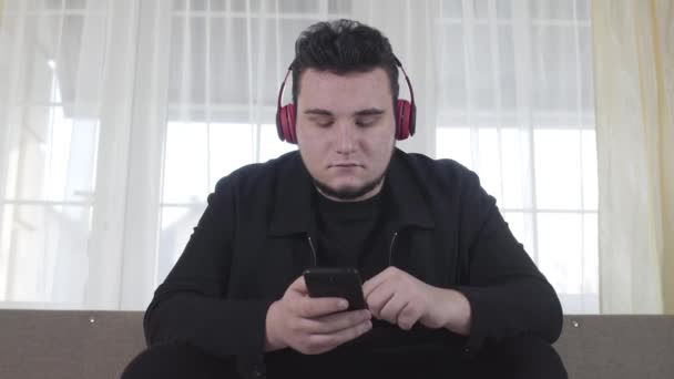 Portrait of plump adult Caucasian man in earphones sitting on couch and using smartphone. Serious guy listening to music in headphones. Lifestyle, hobby, leisure. — Stock Video