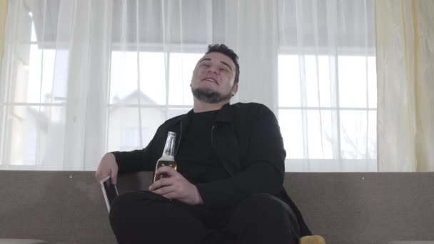 Jeune homme caucasien souriant utilisant une tablette et boire de la bière le week-end. Heureux homme dodu assis sur le canapé et profitant de temps libre à l'intérieur. Mode de vie, loisirs, détente . — Video