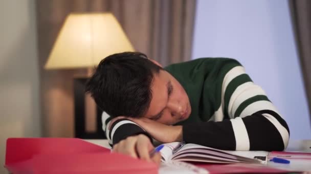 Primer plano del exhausto chico asiático sentado con la cabeza en la mesa. Estudiante universitario cansado tomando un descanso durante el estudio. Sobretrabajo, estudio, concepto educativo . — Vídeos de Stock