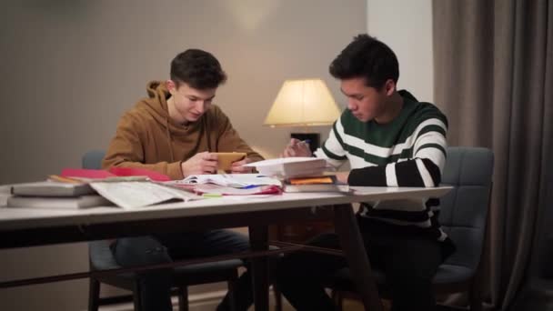 Два студента сидят за столом с книгами и рабочими тетрадями. Азиатский мальчик учится на своего белого друга с помощью смартфона. Образование, домашнее задание, образ жизни . — стоковое видео