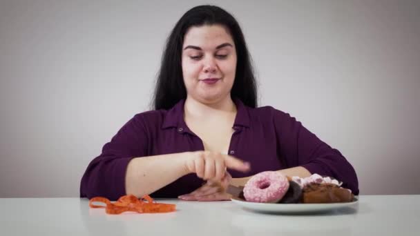 Retrato de menina obesa branca sorridente sentada na frente de doces e fita métrica. Mulher gorda se movendo medidor de fita e tomando prato com sobremesa. Obesidade, dieta, autocontrole . — Vídeo de Stock