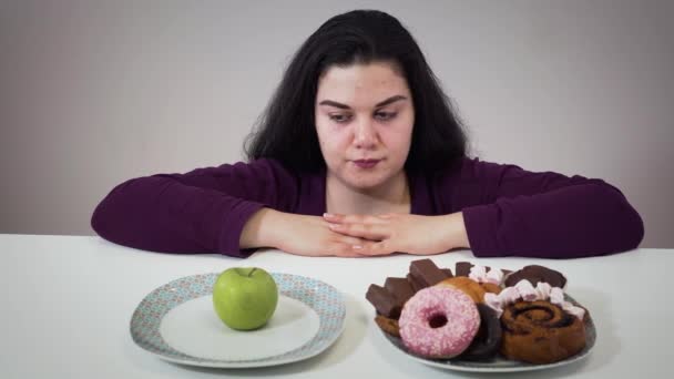 Portrait of thoughtful brunette Caucasian woman looking at plates with apple and sweets. Obese girl selecting between healthy and tasty foods. Overweight problem, obesity, eating. — 비디오