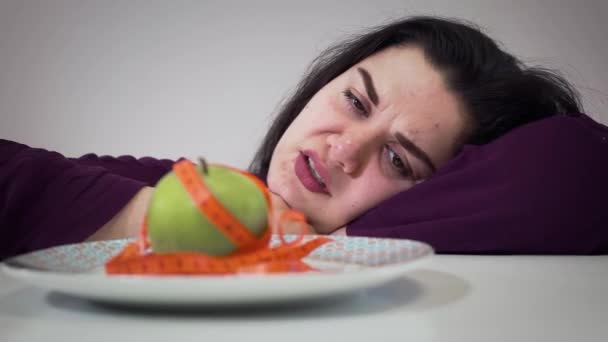 Close-up de triste menina gorda caucasiana olhando com nojo de maçã. Jovem obesa a fazer dieta. Retrato de menina gorda na frente da placa com comida saudável e fita métrica . — Vídeo de Stock