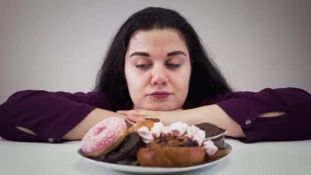 Камера наближається до засмученої товстої кавказької жінки, яка сидить навпроти солодощів і облизуючих губ. Дівчина з ожирінням дивиться на смачний десерт у тарілці. Огрядна вага, переїдання, нездоровий спосіб життя. — стокове відео