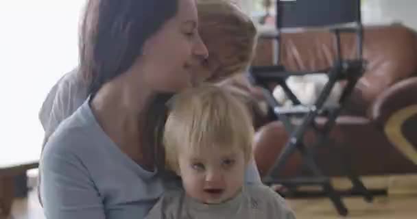 Young Caucasian mother sitting on the floor with baby daughter as her elder son kissing her on cheek. Happy woman spending time with children. Happiness, love, tenderness. Cinema 4k ProRes HQ. — Stock Video