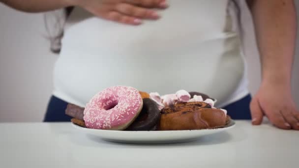 Mulher gorda caucasiana irreconhecível acariciando a barriga, prato com sobremesa em primeiro plano. Menina obesa com comida insalubre. Sobrepeso, estilo de vida insalubre, dieta . — Vídeo de Stock