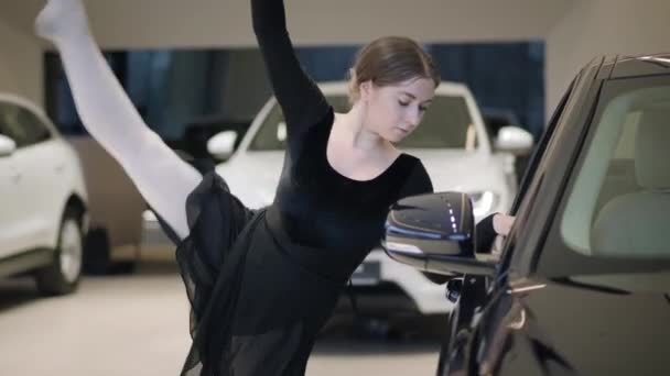 迷人的白人芭蕾舞演员弯腰坐在黑色轿车旁边。漂亮的年轻女子在汽车经销店跳芭蕾舞.汽车陈列室里的芭蕾舞演员.艺术、汽车工业、古典舞蹈. — 图库视频影像