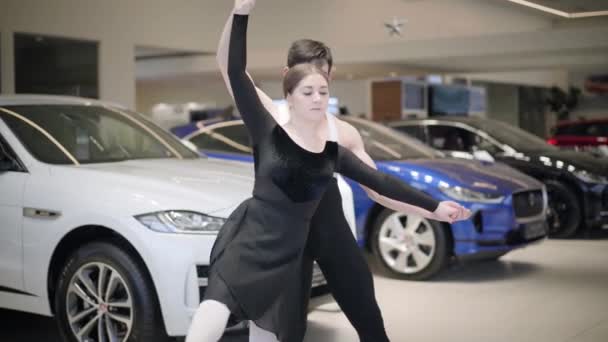 Joven hermosa mujer caucásica girando en un dedo del pie apoyado por el hombre. Bailarines de ballet profesionales bailando en sala de exposición de automóviles. Elegancia, arte, danza clásica . — Vídeos de Stock