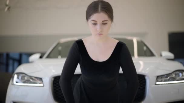 Camera approaching to face of beautiful Caucasian ballerina raising hand up and putting down. Portrait of charming young ballet dancer in car dealership. Elegance, beauty, art. — Stock Video