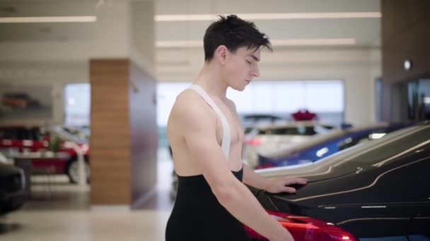 Vista lateral de joven confiado hombre caucásico bailando en concesionario de coches. Guapo bailarín de ballet masculino girando delante de un automóvil negro. Industria automotriz, arte, elegancia . — Vídeos de Stock