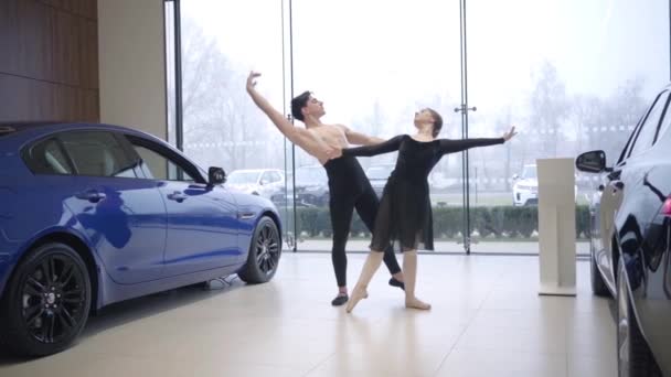 Largo tiro de jóvenes bailarines de ballet caucásicos bailando en concesionarios de automóviles. Hombre y mujer realizando danza clásica en sala de exposición de automóviles. Arte, elegancia, estilo de vida. Movimiento lento . — Vídeos de Stock