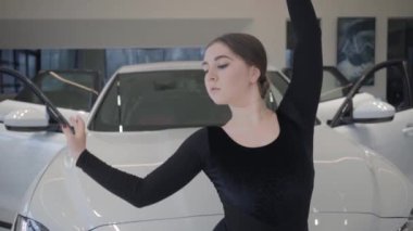 Slowmo of beautiful Caucasian female ballet dancer moving in front of white automobile. Young woman dancing in car dealership. Elegance, lifestyle, art, auto industry. Slow motion.