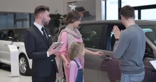 Joven familia caucásica la selección de automóviles nuevos en la sala de exposición de coches. Hombre, mujer y niña linda de pie con concesionario de coches en sala de exposición automática y hablando. Estilo de vida, negocios. Sede del cine 4k ProRes . — Vídeo de stock