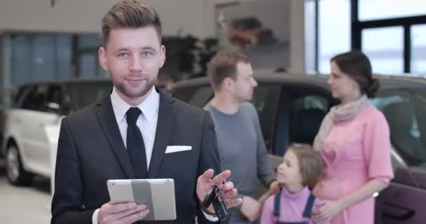 Retrato de jovem confiante negociante de carro masculino segurando tablet e chaves do carro, olhando para a câmera. Família caucasiana jovem conversando no fundo. Comerciante profissional vendendo automóvel. Cinema 4k ProRes HQ . — Vídeo de Stock