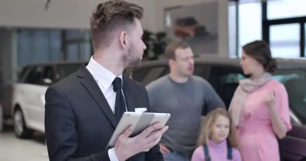 Jovem caucasiano sorridente olhando para a família conversando no fundo e mostrando as chaves do carro para a câmera. Vendedor masculino positivo que vende o automóvel aos clientes. Indústria automóvel. Cinema 4k ProRes HQ . — Vídeo de Stock