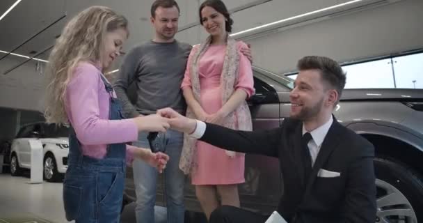 Λευκός άντρας δίνει κλειδιά αυτοκινήτου σε όμορφη ξανθιά κοπέλα και χαμογελάει. χαρούμενο παιδί που κοιτάζει τους γονείς του και πηδάει πάνω. Επιτυχημένη νεαρή οικογένεια που αγοράζει νέο όχημα στην αντιπροσωπεία. Κινηματογράφος 4k Prores Hq. — Αρχείο Βίντεο