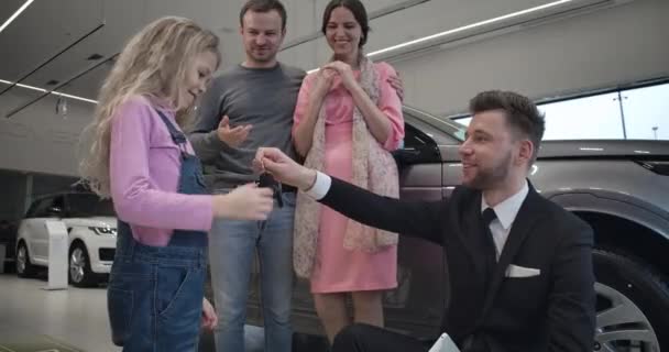 男性ディーラーから車の鍵を取っているかなり小さな白人の女の子は、父親と抱擁の両親にそれらを与えます。自動車販売の新しい自動車を購入する若い家族を満足させました。Cinema 4k Prores｜Hq. — ストック動画