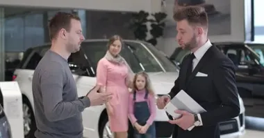 Kafkasyalı genç işadamı arabanın anahtarlarını konuşuyor ve arka planda mutlu karısı ve kızı kucaklaşırken tüccarların elini sıkıyor. Başarılı bir adam galeriden yeni bir araba alıyor. Sinema 4k Prores Merkezi.