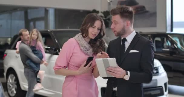 Pewna siebie biała bizneswoman rozmawia z dealerem samochodów jako jej mąż i córka stojący w tle. Elegancka kobieta kupująca nowy samochód w salonie samochodowym. Kino 4k Prores Hq. — Wideo stockowe