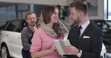 Elinde küçük tatlı bir kızla gülümseyen beyaz bir adam arka plandan gelen başarılı bir kadınla torbacı ile konuşuyor. Araba galerisinde genç bir aile otomobil satın alıyor. Sinema 4k Prores Merkezi.