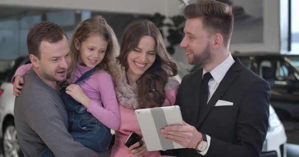 Close-up portrait of four Caucasian people standing in car dealership and talking. Smiling young family discussing automobile purchase with trader in showroom. Auto industry. Cinema 4k ProRes HQ. — 비디오