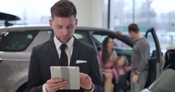 Portrait of confident young Caucasian man using tablet as family talking in car at the background. Successful car dealer selling automobile and gesturing yes by shaking head. Cinema 4k ProRes HQ. — Stock Video